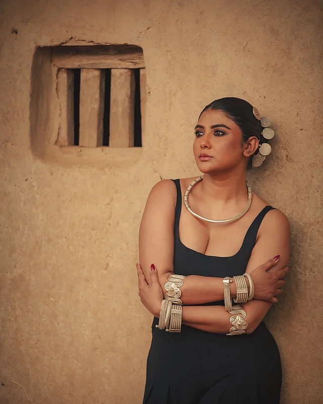 অভিনেত্রীর পরনে স্লিভলেস কালো জাম্পস্যুট আর ট্রাইবাল গয়না, যা একদিকে আধুনিক মিনিমাল লুক দিচ্ছে, অন্যদিকে গ্রামীণ মাটির দেয়ালের পটভূমিতে কনট্রাস্ট তৈরি করছে।