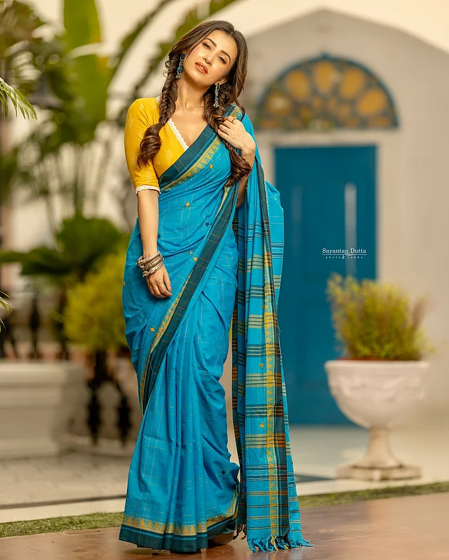 মনামী পরেছেন নীল তাঁতের শাড়ি। শাড়ির জমিন ও পাড়ে থাকা নকশার সঙ্গে মিলিয়ে সাদা লেইস দেওয়া হলুদ ব্লাউজ পরেছেন সঙ্গে।
