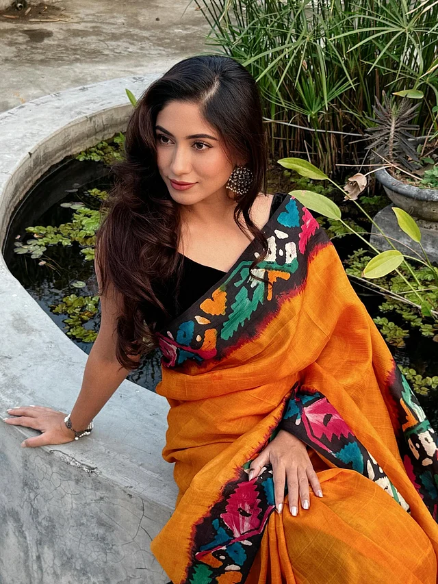 প্রথমেই নজর কাড়ছে শাড়ির রঙের দাপট। উজ্জ্বল কমলা আর কালোর কনট্রাস্টের ওপর জ্যামিতিক বহুরঙা নকশা খেলা করছে। জুটি হয়েছে ভেলভেটের কালো ব্লাউজ।