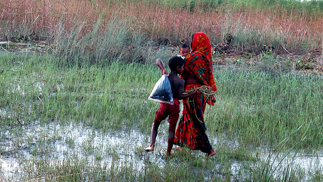 মাঠের পানি মাড়িয়ে সন্তানদের নিয়ে বাড়ি যাচ্ছেন এক মা। ছবি: নেয়ামতউল্যাহ, ভোলা
