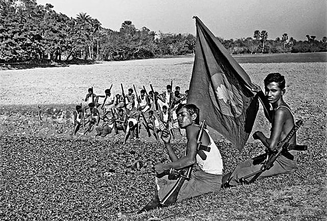 পতাকা হাতে এক মুক্তিযোদ্ধা মুখ দিয়ে আঁচড় কাটছেন ঘাসফুলে, পাশে আরেক মুক্তিযোদ্ধা। ছবি: আনোয়ার হোসেন, দোহার, ১৯৭১