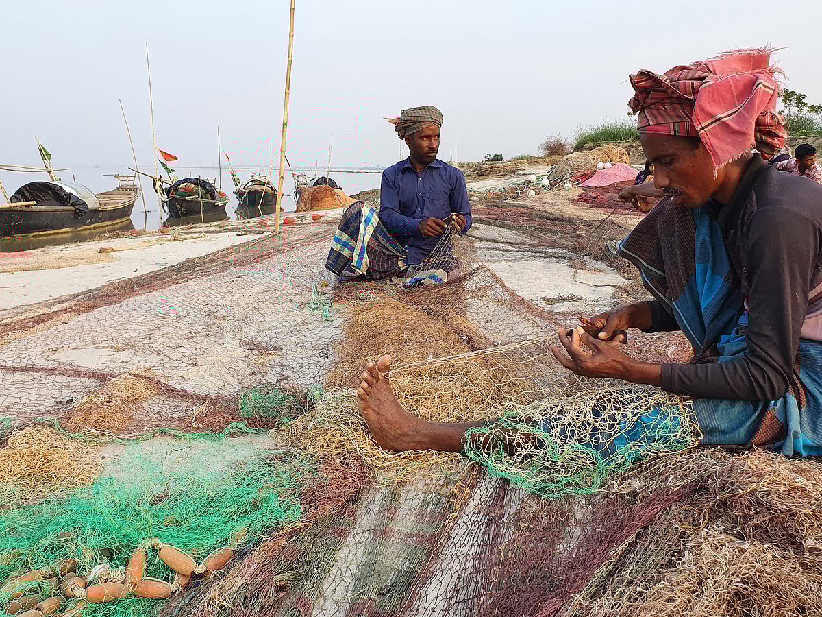 নদীর পাড়ে বসেই জাল মেরামত করছেন জেলেরা।
