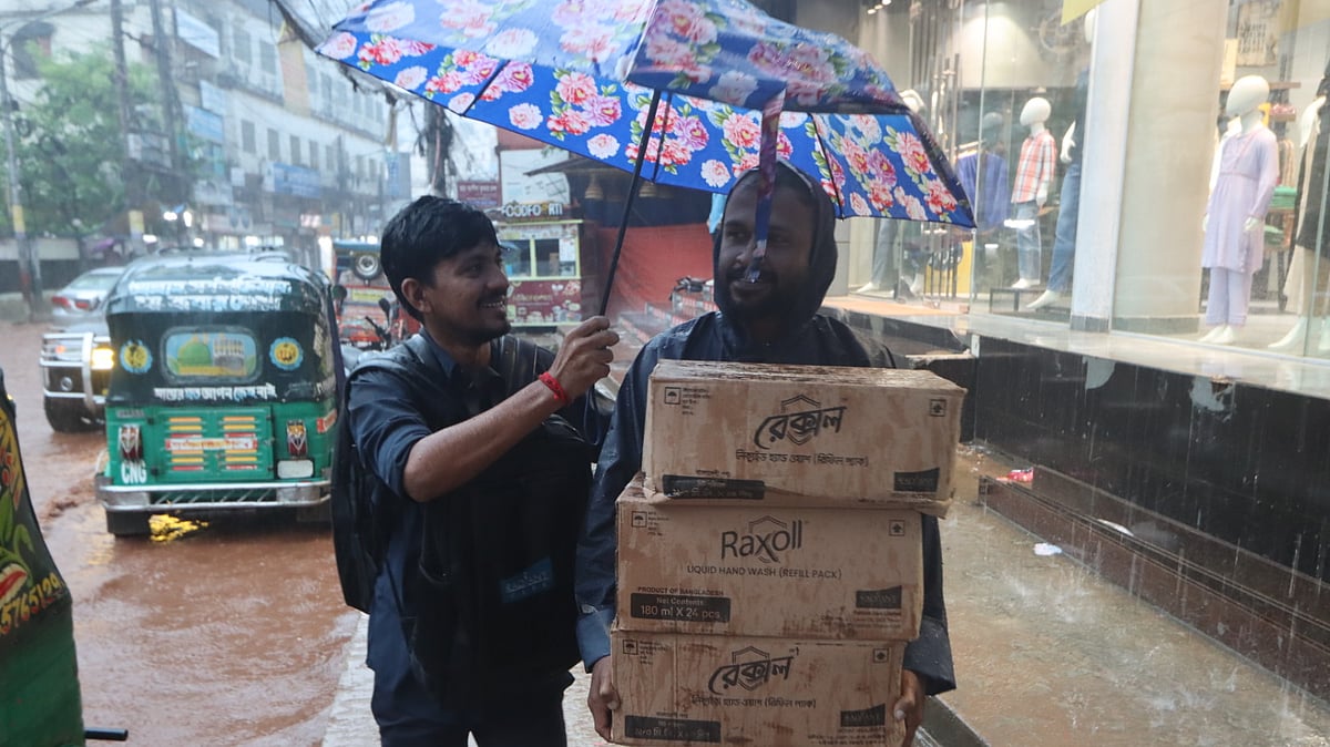 বৃষ্টির মধ্যে ছাতা মাথায় দিয়ে পণ্য দোকানে নিয়ে যাচ্ছেন দুজন বিক্রয় কর্মী।