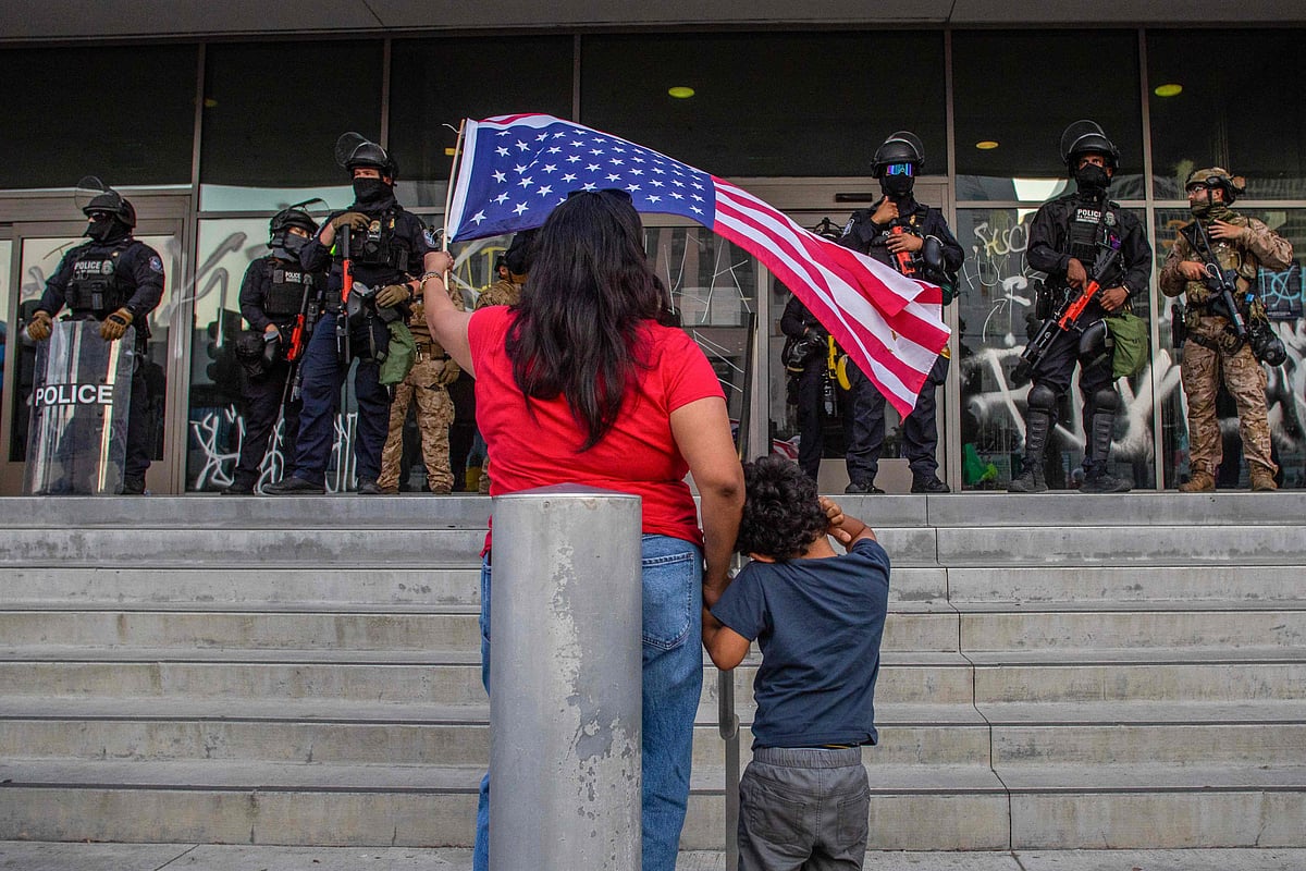 এক নারী এক হাতে একটি শিশুর হাত ধরে অন্য হাতে যুক্তরাষ্ট্রের পতাকা নাড়াচ্ছেন। এ সময় লস অ্যাঞ্জেলেসে ফেডারেল ভবনের বাইরে দাঁড়িয়ে আছেন ক্যালিফোর্নিয়া ন্যাশনাল গার্ডের সদস্যরা। ১০ জুন ২০২৫