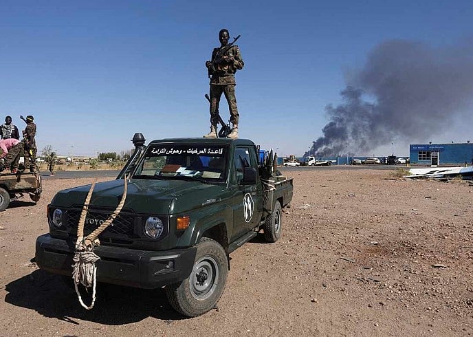 গাড়ির ওপরে দাঁড়িয়ে আছেন সুদানের সেনাবাহিনীর সদস্য। পেছনে জ্বলছে তেল পরিশোধনাগার। সুদানের উত্তর বাহরি প্রদেশ। জানুয়ারি, ২০২৫