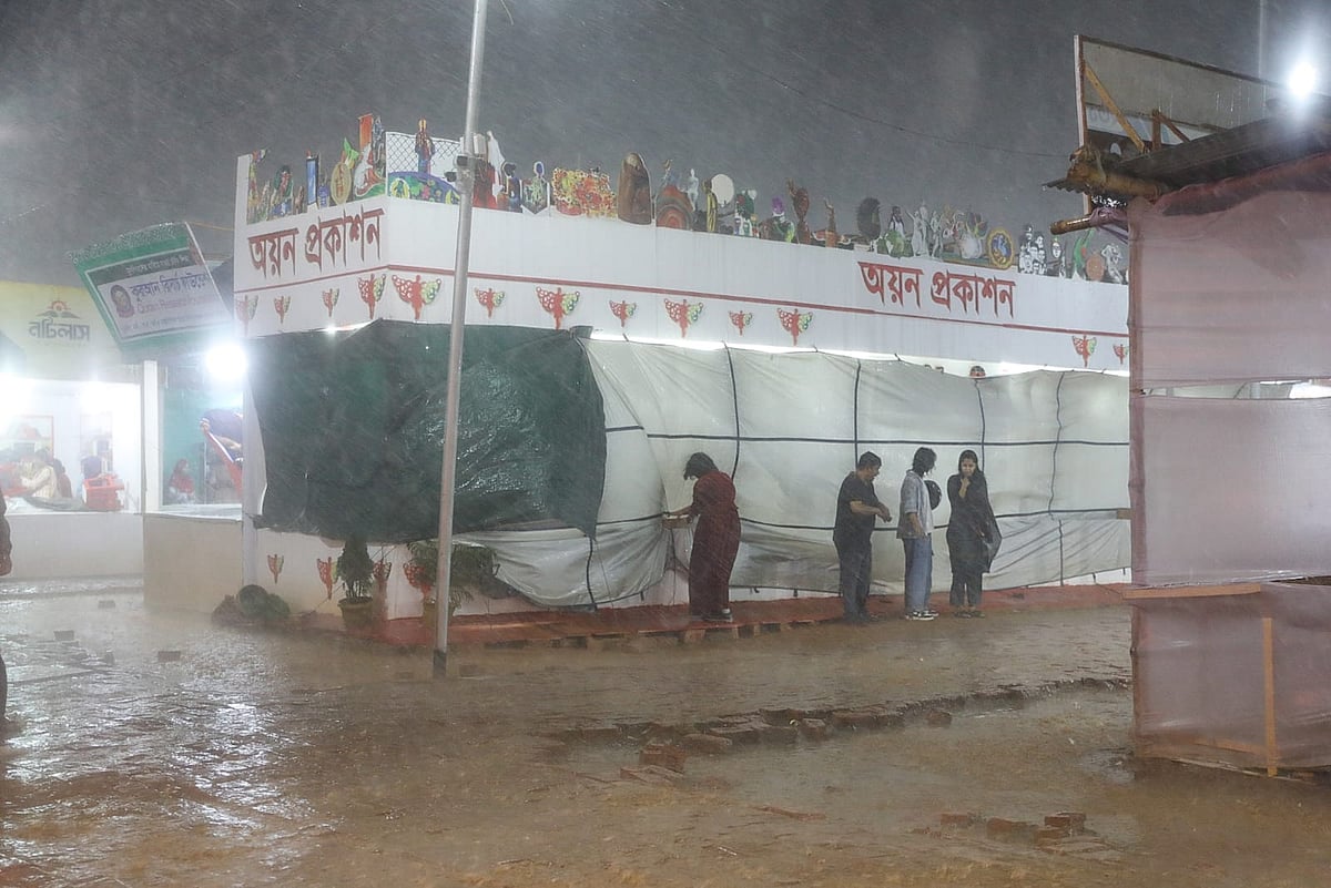 বৃষ্টির মধ্যে বইয়ের স্টলের পাশে দাঁড়িয়ে ভিজছেন কয়েকজন। অমর একুশে বইমেলা, ১৩ মার্চ ২০২৬