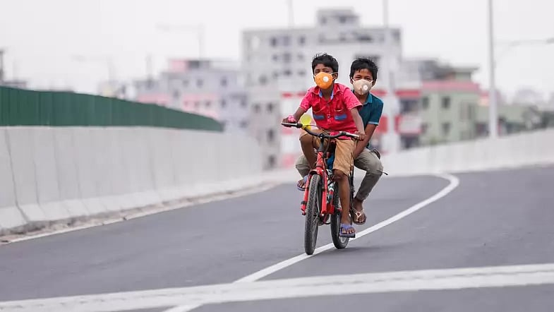 Two children wearing face masks ride a bicycle on a flyover.