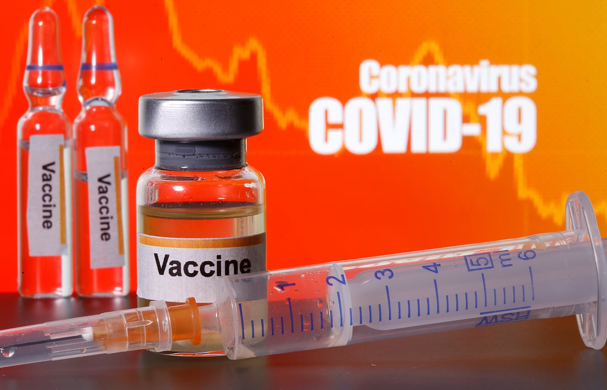 Small bottles labeled with "Vaccine" stickers stand near a medical syringe in front of displayed "Coronavirus COVID-19" words in this illustration.