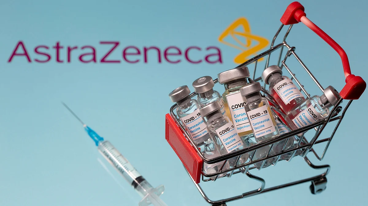 A small shopping basket filled with vials labeled "COVID-19 - Coronavirus Vaccine" and a medical sryinge are placed on a AstraZeneca logo