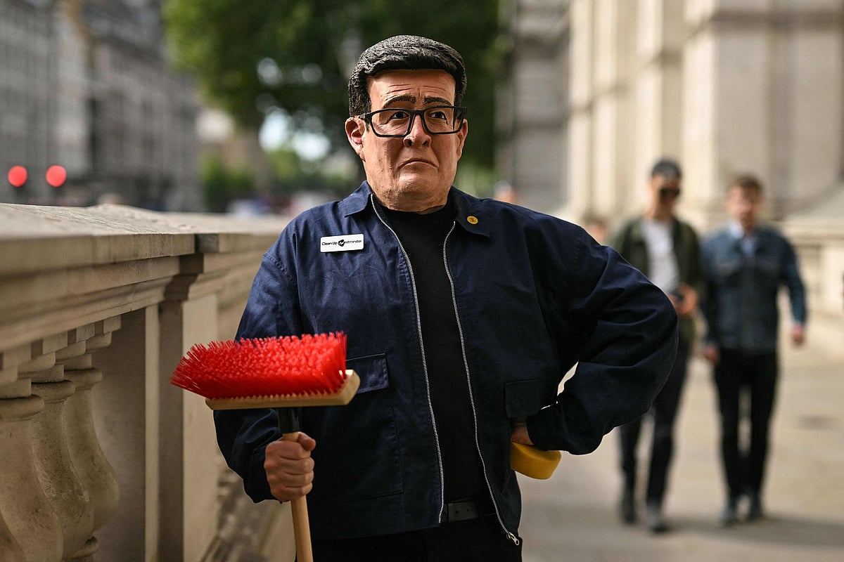 A man wearing a mask of Britain's incoming Prime Minister Andy Burnham holds a broom while standing on the pavement outside Downing Street in central London on 20 July 2026, ahead of Burnham's arrival as the country's new leader.